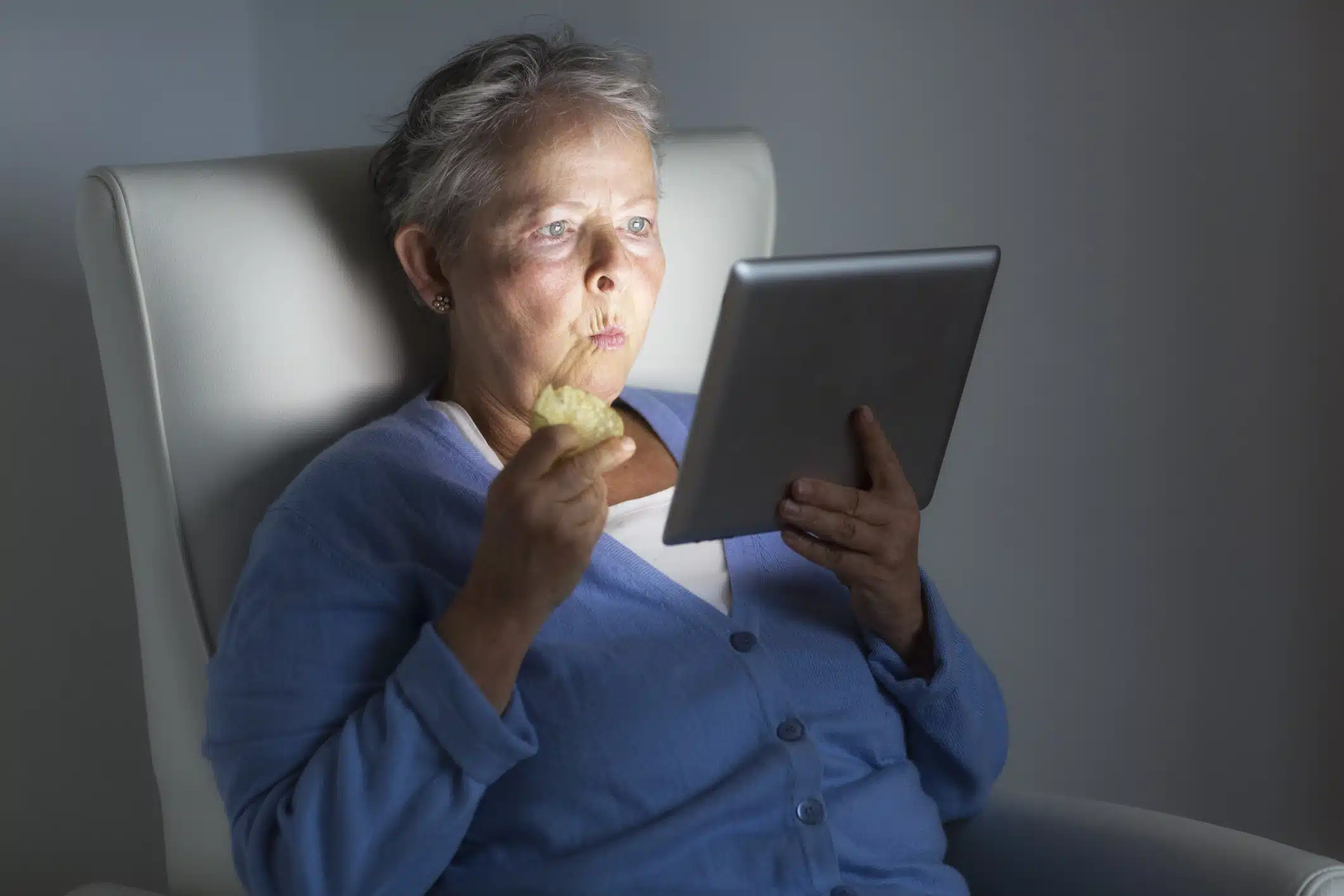 Donna anziana che mangia e guarda il tablet seduta su una poltrona